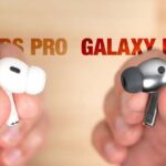 A close-up comparison image showing white Apple-style wireless earbuds on the left labeled “AirPods Pro” and gray Samsung-style earbuds on the right labeled “Galaxy Buds3,” both held between fingers against a blurred background.