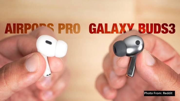 A close-up comparison image showing white Apple-style wireless earbuds on the left labeled “AirPods Pro” and gray Samsung-style earbuds on the right labeled “Galaxy Buds3,” both held between fingers against a blurred background.